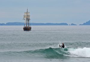 A surfer hops off the lip of a shoulder-high wave as a tallship cruises towards the harbor in the background