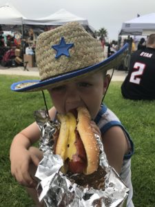 A toddler in a cowboy hat takes a huge bite of hotdog