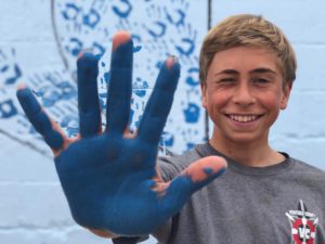 A smiling teen holds the blue paint-covered palm of his hand up for the camera