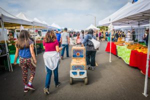 A vendor pulls a cart full of Food Forward produce past Farmers Market visitors