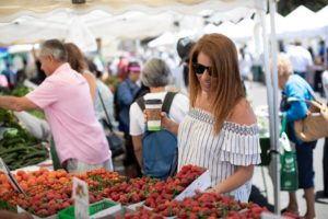 A smiling woman with coffee in hand, buying strawberries at the Farmers Market