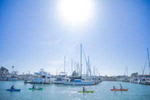 A group of kayaks paddles past boats moored in Channel Islands Harbor