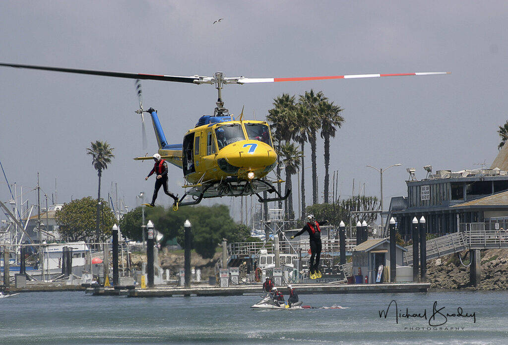 A yellow helicopter hovers over water with a rescuer rappelling down, while another is lifted up. A jet ski is nearby, and boats are visible in the background.