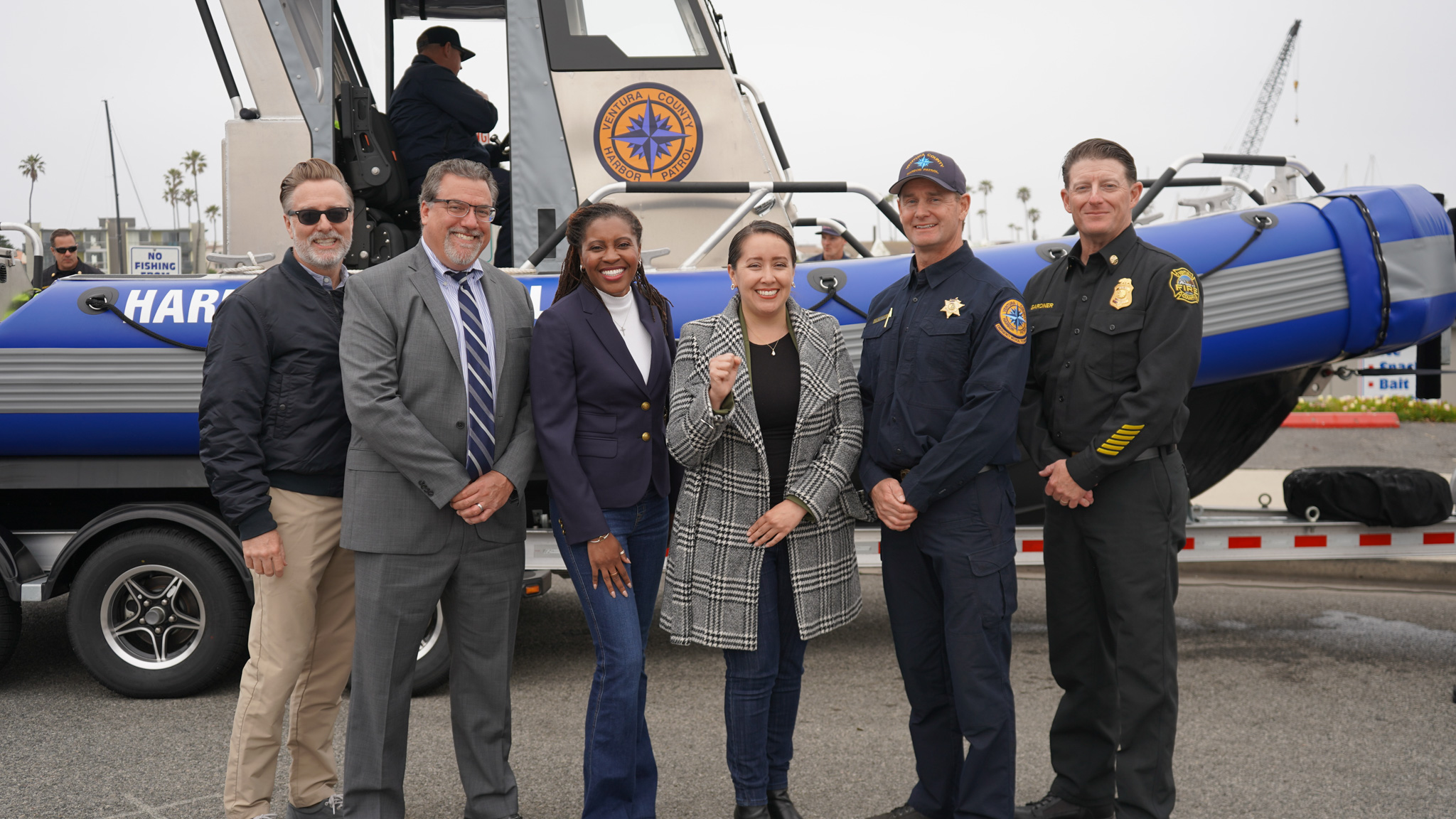 Six people stand in front of a blue rescue boat on a trailer, posing for a group photo; some are in uniforms and others in business attire.