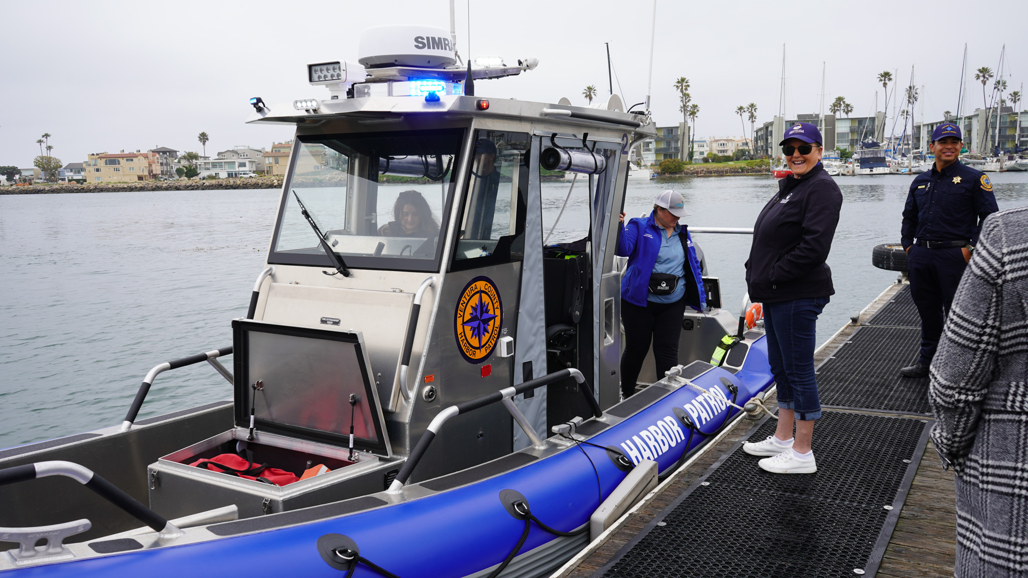 People stand on a dock next to a harbor patrol boat, with one person onboard and others nearby; buildings and palm trees are visible in the background.
