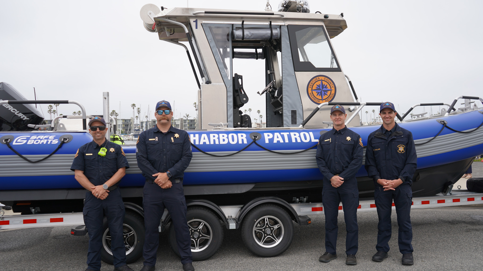 Four uniformed harbor patrol officers stand in front of a blue and gray patrol boat on a trailer.