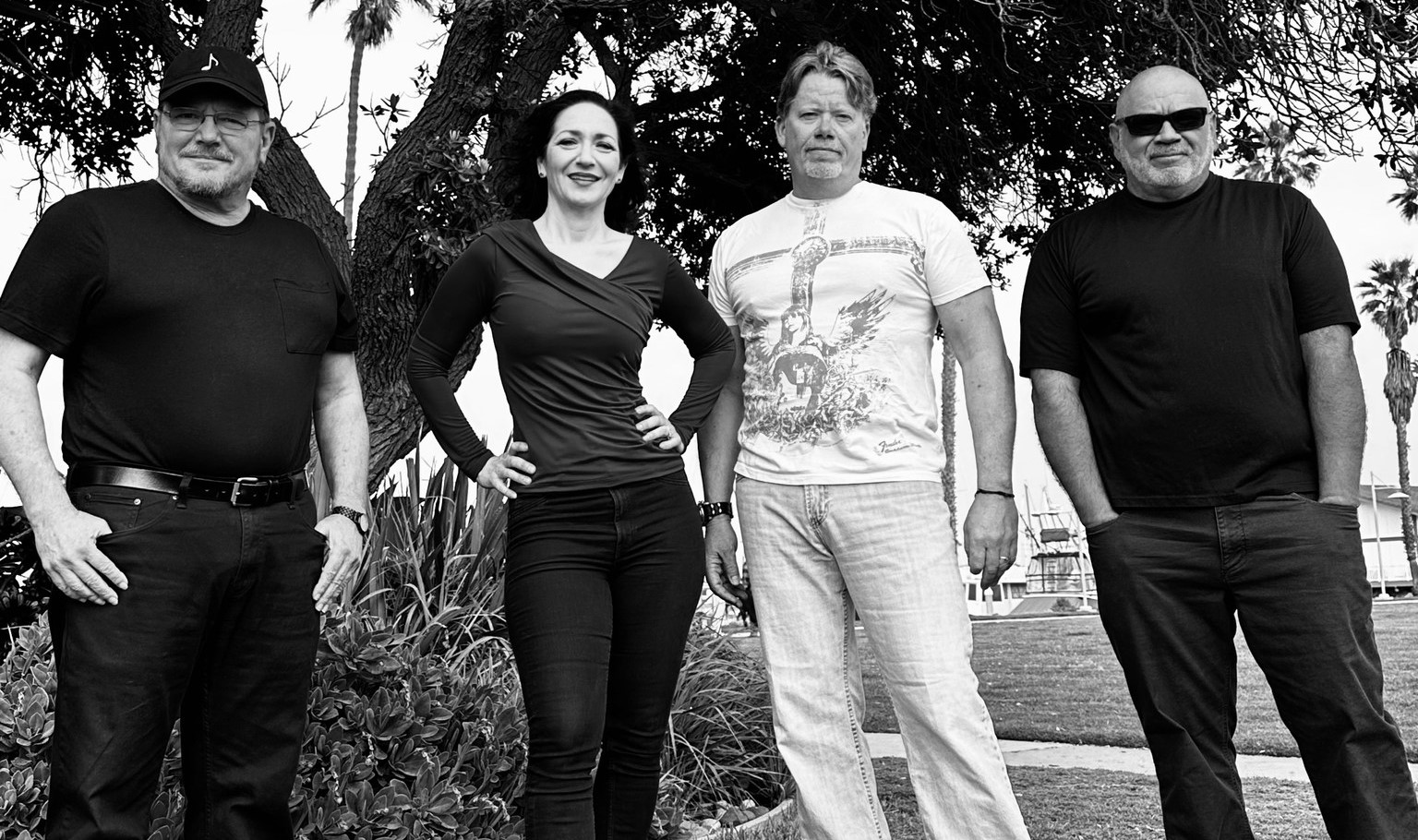 Four adults stand outdoors in front of trees and greenery, posing for a group photo on a sunny day.
