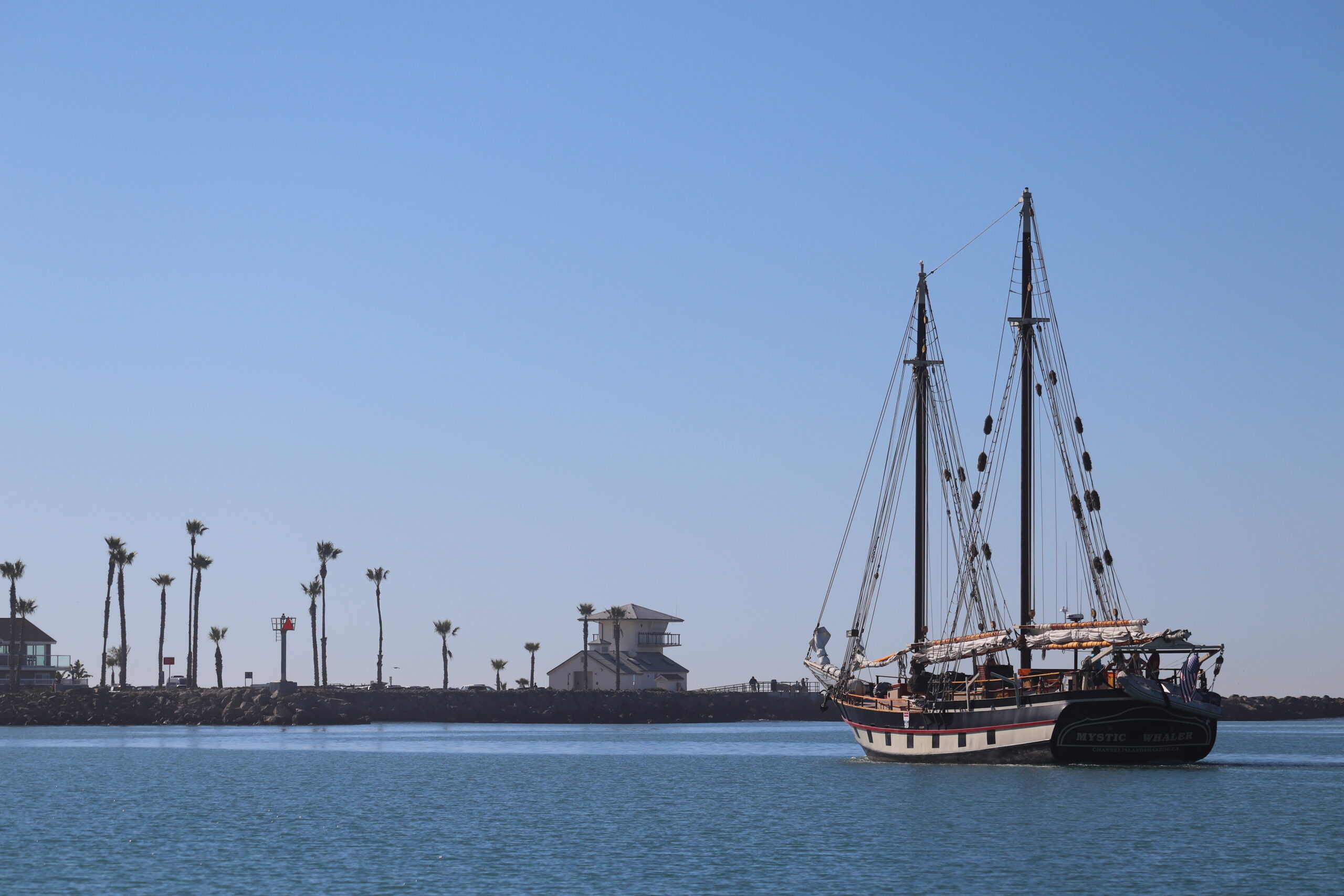 A two-masted sailing ship floats on calm water near a shoreline with palm trees and buildings under a clear blue sky.