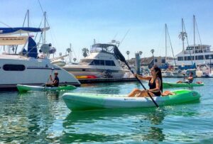 Three people kayak near docked boats and yachts in a marina on a sunny day.